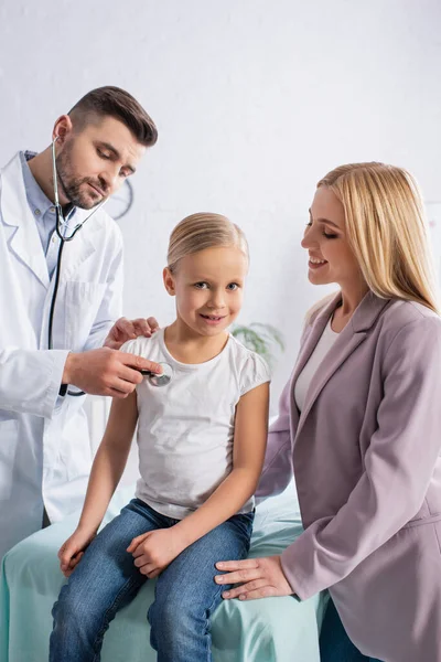 Улыбающийся ребенок смотрит в камеру рядом с мамой и доктором со стетоскопом — стоковое фото