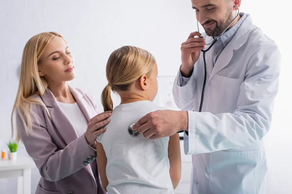 Médico sonriente con estetoscopio examinando la espalda del niño cerca de los padres - foto de stock