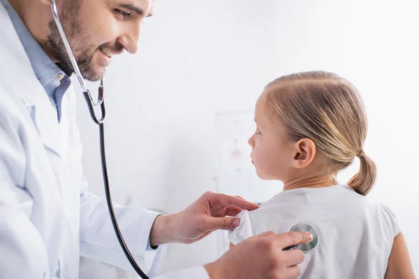 Médico sonriente examinando a un niño con estetoscopio en la clínica - foto de stock