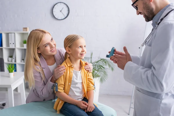 Pediatra sosteniendo inhalador cerca de niño y mamá en el hospital - foto de stock