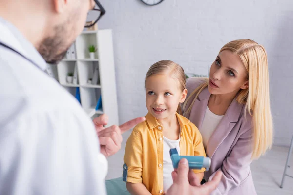 Жінка дивиться на сімейного лікаря, вказуючи на інгалятор біля усміхненої дочки — стокове фото