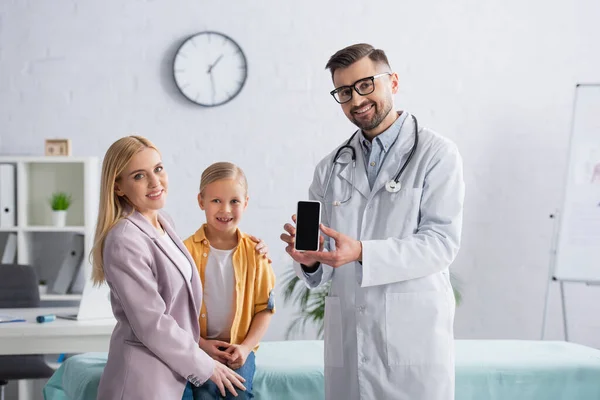 Positive pediatrician holding smartphone with blank screen near family in clinic — Stock Photo