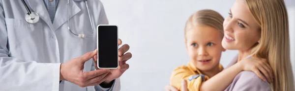 Doctor sosteniendo teléfono inteligente con pantalla en blanco cerca de la madre y el niño sonrientes, pancarta - foto de stock