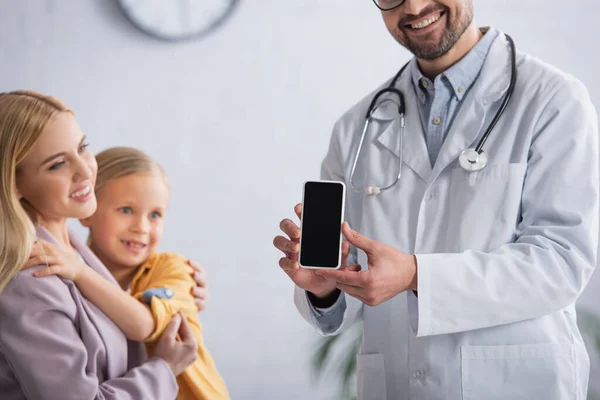 Smiling family doctor holding cellphone near family on blurred background in clinic — Stock Photo