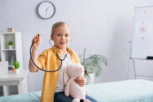 Excited child in stethoscope holding soft toy on medical couch in hospital — Stock Photo