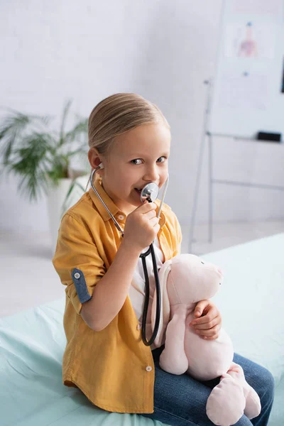 Smiling kid playing with soft toy and stethoscope on medical couch — Stock Photo