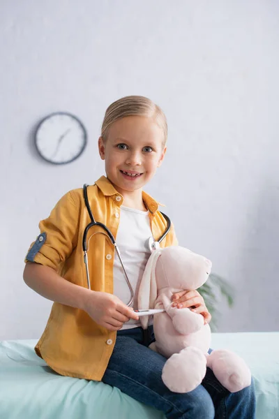Smiling kid looking at camera while holding soft toy and electronic thermometer — Stock Photo