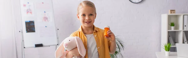 Criança sorridente segurando brinquedo macio e pílulas na clínica, banner — Fotografia de Stock