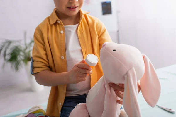 Vue recadrée d'un enfant tenant des pilules près d'un jouet doux et d'un thermomètre flou à la clinique — Photo de stock