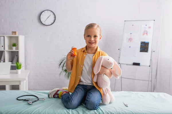 Mädchen mit Pillen und Stofftier lächelt in die Kamera neben Stethoskop und elektronischem Thermometer — Stockfoto