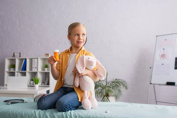 Child smiling and holding pills with soft toy on medical couch in hospital — Stock Photo