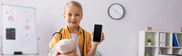 Fille souriante avec lapin jouet tenant téléphone portable avec écran blanc à la clinique, bannière — Photo de stock