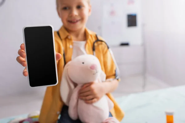 Chica borrosa con juguete suave que muestra el teléfono móvil con pantalla en blanco en el hospital - foto de stock