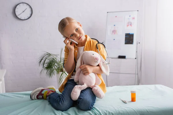 Criança com coelho de brinquedo sentado no sofá médico e falando no smartphone — Fotografia de Stock