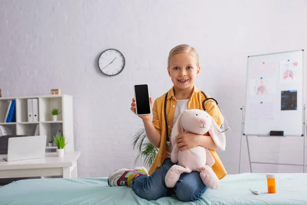 Fille heureuse assise sur le canapé médical avec lapin jouet et montrant smartphone avec écran vide — Photo de stock