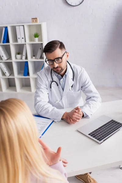 Doctor sitting near laptop and clipboard while listening to blurred woman — Stock Photo