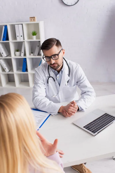 Médico sentado cerca de la computadora portátil y portapapeles mientras escucha a la mujer borrosa - foto de stock