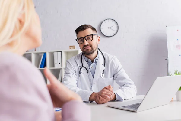 Doctor in white coat and eyeglasses listening to blurred patient in clinic — Stock Photo