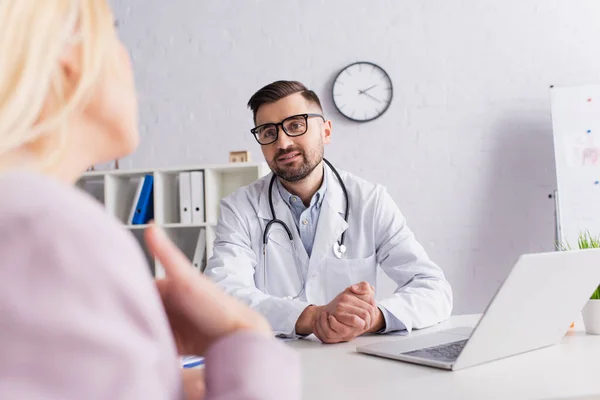 Médecin en manteau blanc et lunettes écoutant patient flou à la clinique — Photo de stock