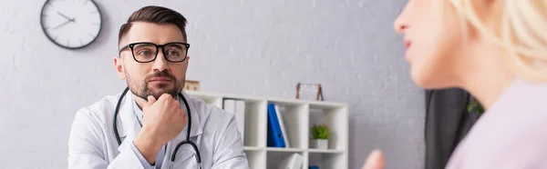 Médico segurando a mão perto do queixo enquanto ouve mulher turva, banner — Fotografia de Stock