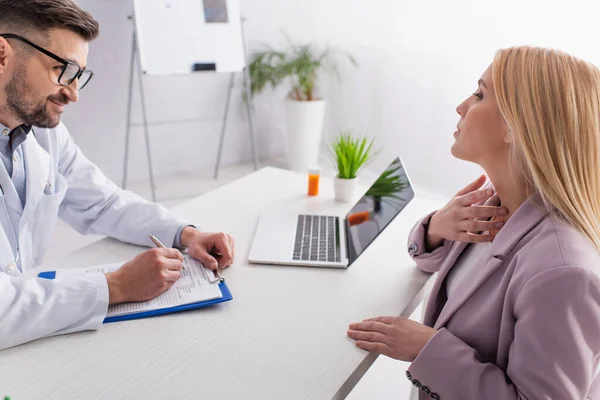 Smiling doctor writing prescription near woman touching sore throat during consultation — Stock Photo