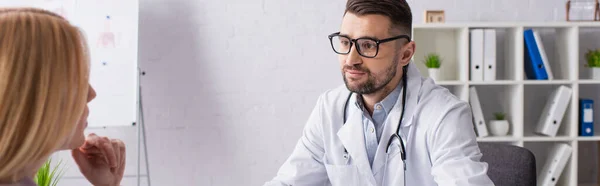 Doctor en bata blanca y anteojos cerca de paciente rubio en clínica, pancarta - foto de stock