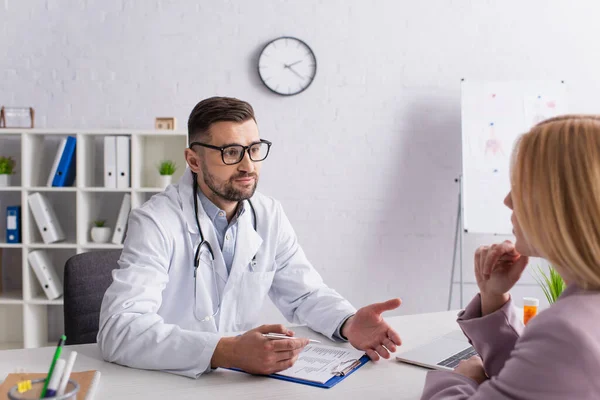 Doctor in white coat talking to blurred woman near clipboard with diagnosis — Stock Photo