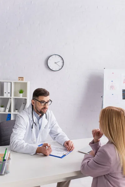 Doctor en bata blanca hablando con mujer rubia cerca del portapapeles con receta - foto de stock