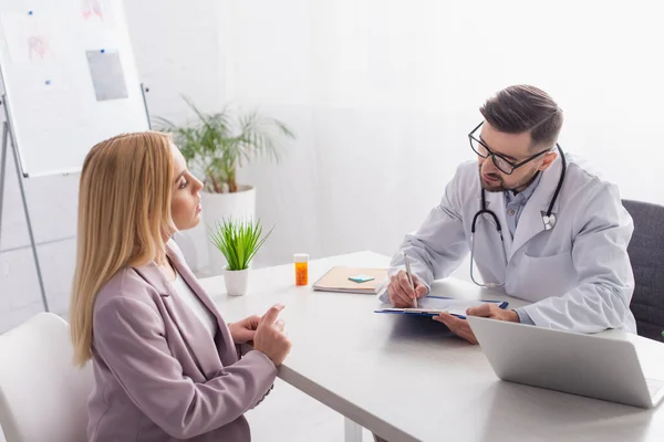 Doctor writing prescription near blonde woman, laptop and container with pills — Stock Photo