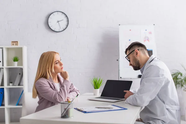 Médecin pointant vers ordinateur portable avec écran vierge près femme blonde à la clinique — Photo de stock