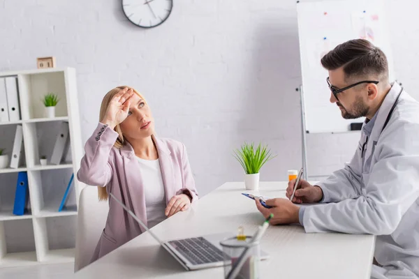 Médecin écrivant le diagnostic tout en regardant la femme tenant la tête près du front pendant la consultation — Photo de stock