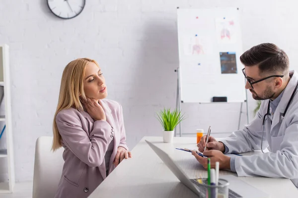 Femme touchant le cou pendant la consultation avec le médecin écrivant sur le presse-papiers — Photo de stock