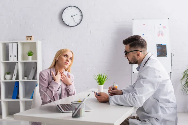 Arzt schreibt auf Klemmbrett, während er Frau ansieht, die während der Konsultation Halsschmerzen berührt — Stockfoto