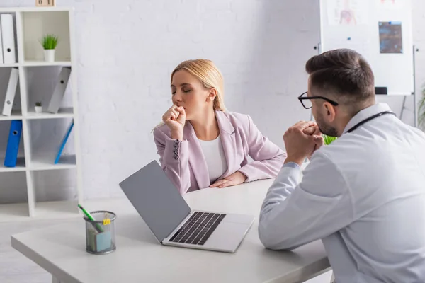 Doctor sitting near laptop with blank screen while looking at woman coughing during consultation — Stock Photo