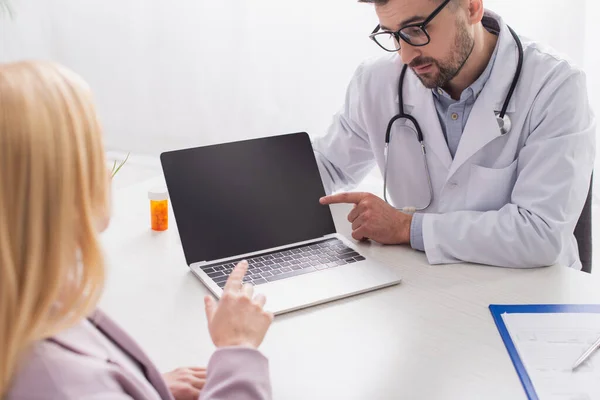 Doctor and blurred woman pointing at laptop during conversation in consulting room — Stock Photo