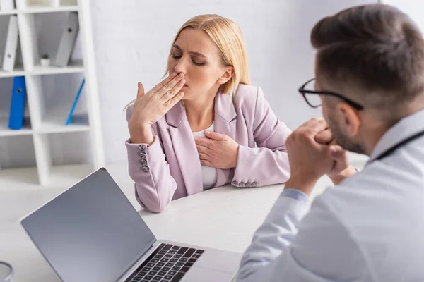 Woman coughing near blurred physician and laptop with blank screen in consulting room — Stock Photo