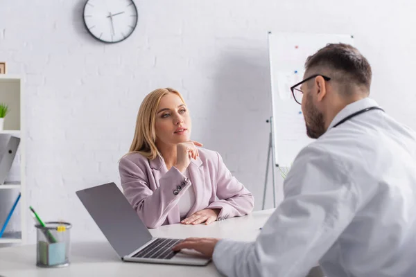 Doctor and blonde woman talking in consulting room near laptop with blank screen — Stock Photo