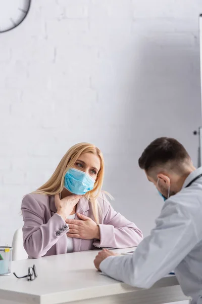 Kranke Frau in medizinischer Maske berührt Brust und Rachen in der Nähe von Arzt — Stockfoto