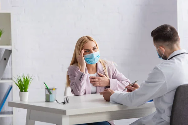 Blonde woman in protective mask suffering from headache near physician — Stock Photo