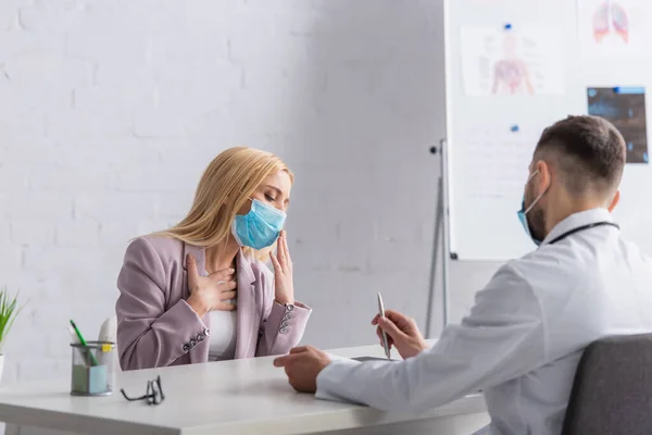 Mujer enferma con máscara médica tosiendo y tocando el pecho cerca del médico - foto de stock