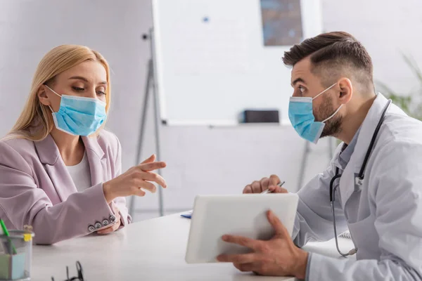 Médico e paciente em máscaras médicas apontando para comprimido digital durante a consulta — Fotografia de Stock