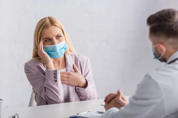Kranke Frau in medizinischer Maske berührt Kopf und Brust bei Konsultation mit verschwommenem Arzt — Stockfoto