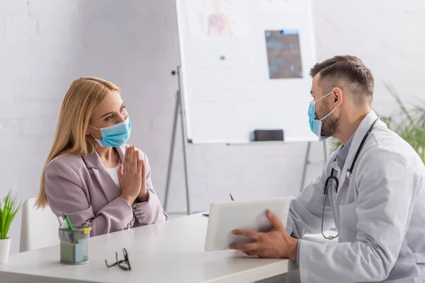 Mujer en máscara protectora que muestra por favor gesto cerca del médico con la tableta digital - foto de stock