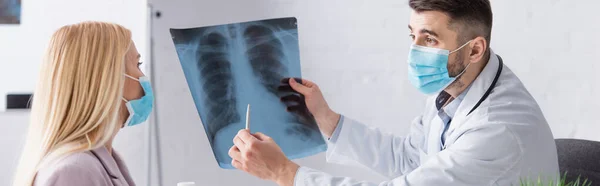 Doctor en máscara médica apuntando a la radiografía de pulmones cerca del paciente, estandarte - foto de stock