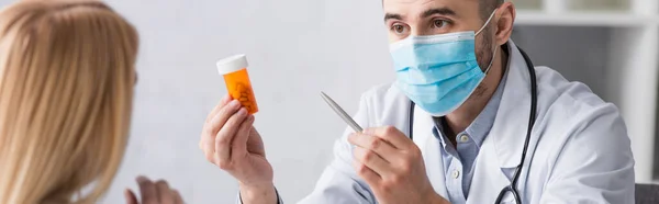 Medico in maschera medica che mostra contenitore con pillole alla donna offuscata, banner — Foto stock