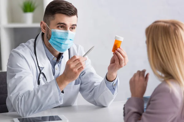 Femme floue près du médecin dans le masque médical pointant vers le récipient avec des pilules — Photo de stock