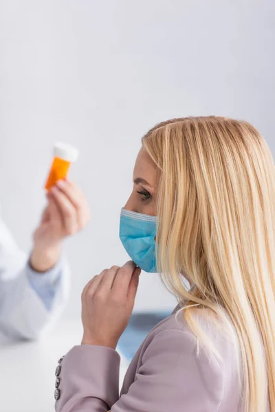 Mujer rubia en máscara médica cerca del médico con pastillas contenedor - foto de stock