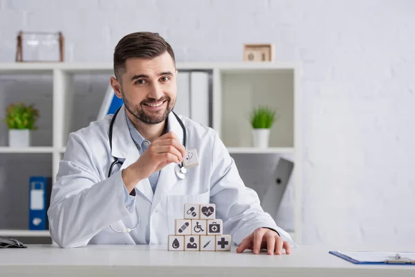 Веселый доктор в белом халате, расставляющий кубики с медицинскими символами в пирамиде на столе — стоковое фото
