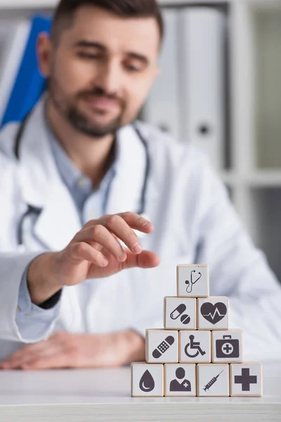 Blurred doctor in white coat near cubes with medical symbols on desk at workplace — Stock Photo