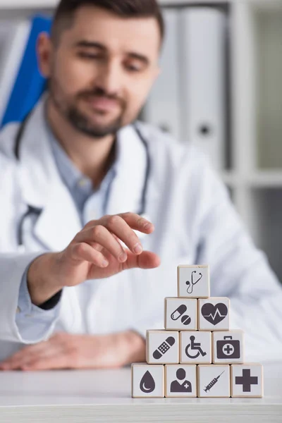 Medico sfocato in cappotto bianco vicino a cubi con simboli medici sulla scrivania sul posto di lavoro — Foto stock